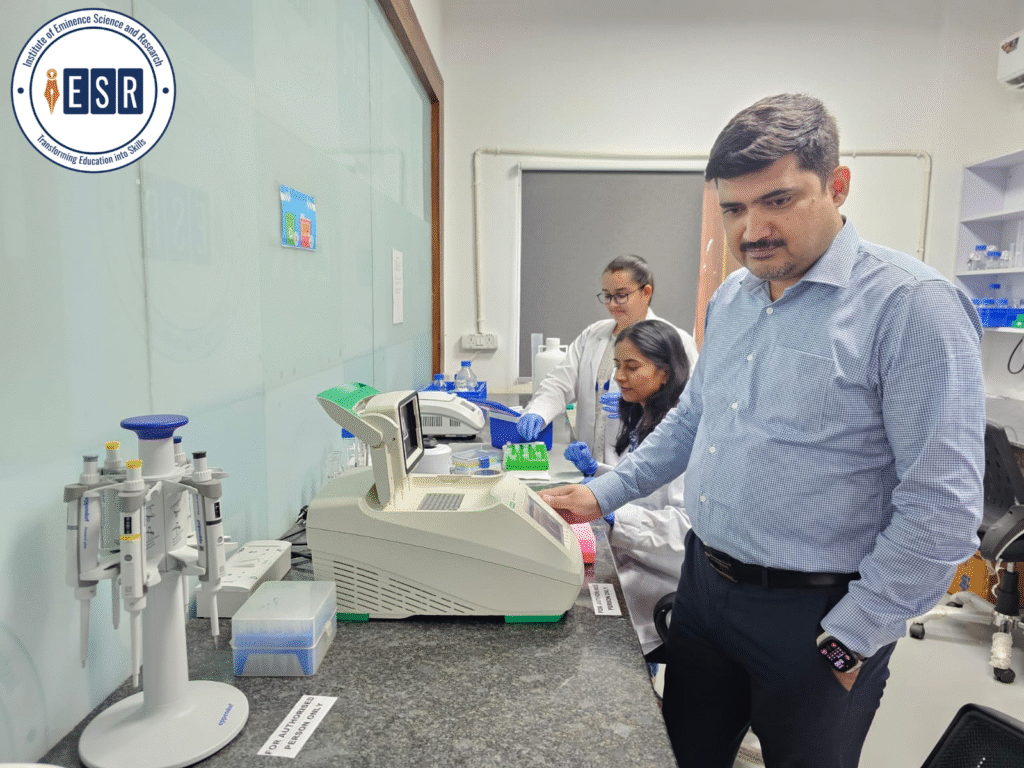 A man in a checkered blue shirt and black pants operates a piece of lab equipment, possibly a plate reader or thermocycler. Two female lab technicians in white coats and gloves stand behind him, observing the process. The lab is clean and modern, with various scientific instruments and a logo for the 'Institute of Eminence Science and Research' visible in the top left corner.